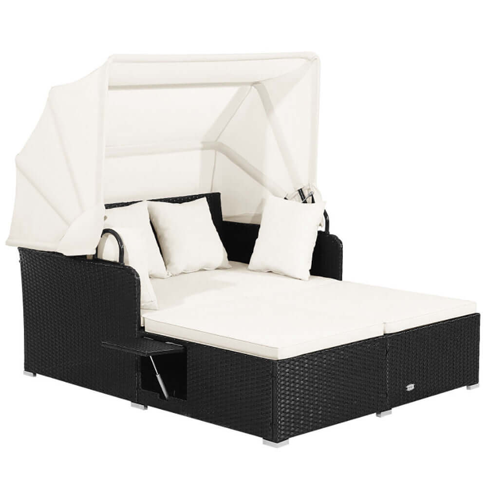 Patio rattan daybed with retractable canopy and cream cushions, featuring side tables for comfort.