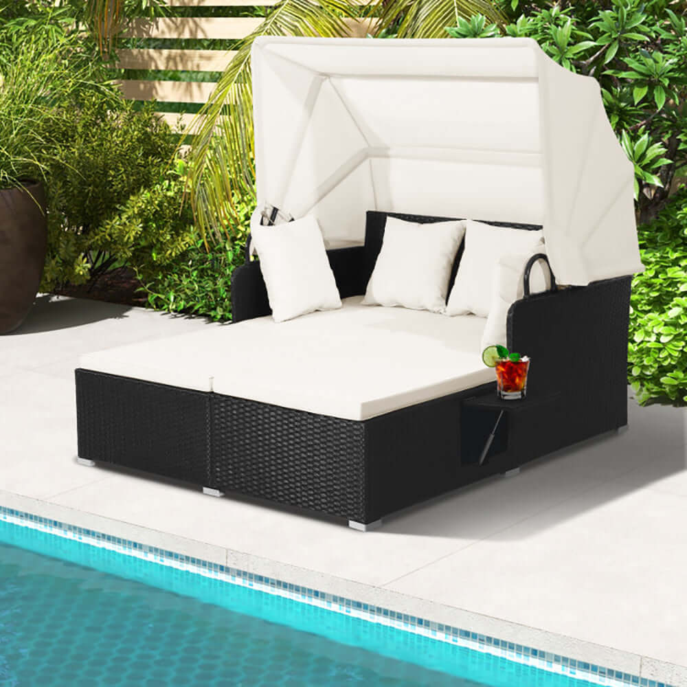 Patio rattan daybed with retractable canopy and cushions near a pool, features side table and tropical greenery.
