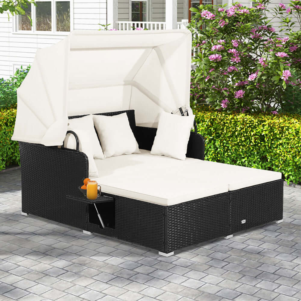 Black patio rattan daybed with retractable canopy and cream cushions in a garden setting.