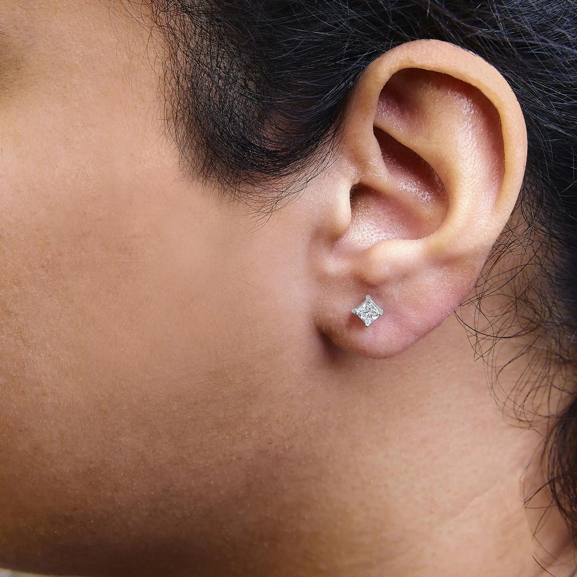 Close-up of a person's ear showcasing 14K white gold princess cut diamond solitaire stud earrings.