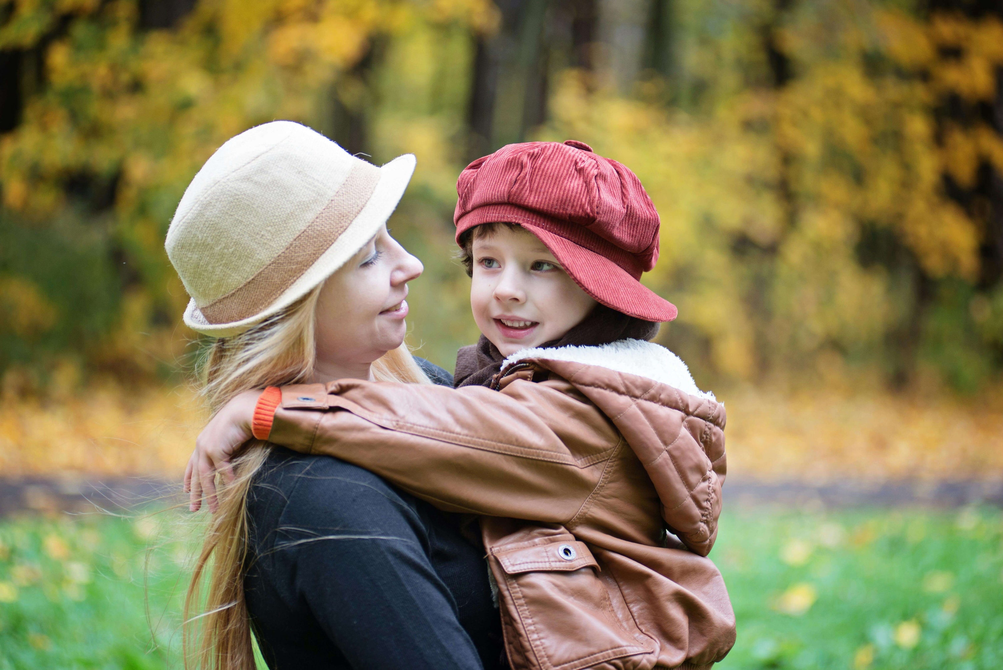 A mother holds her smiling son in a cozy outdoor setting with autumn leaves in the background.